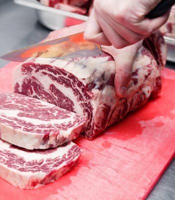 Closeup cook's hand cuts ribeye marbled beef steak with sharp knife on red plastic cutting board on metal table in restaurant kitchen. Concept steakhouse specializing in grilled meat, live fire